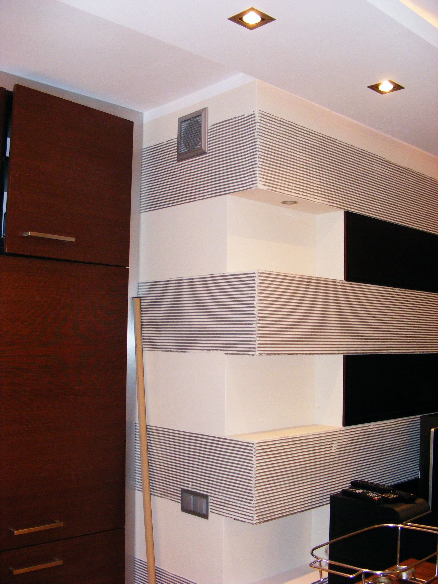 Wykończony narożnik pokoju z półkami, pokryty szpachlą i pomalowany w poziome paski, widoczny fragment szafy i listwa przypodłogowa.